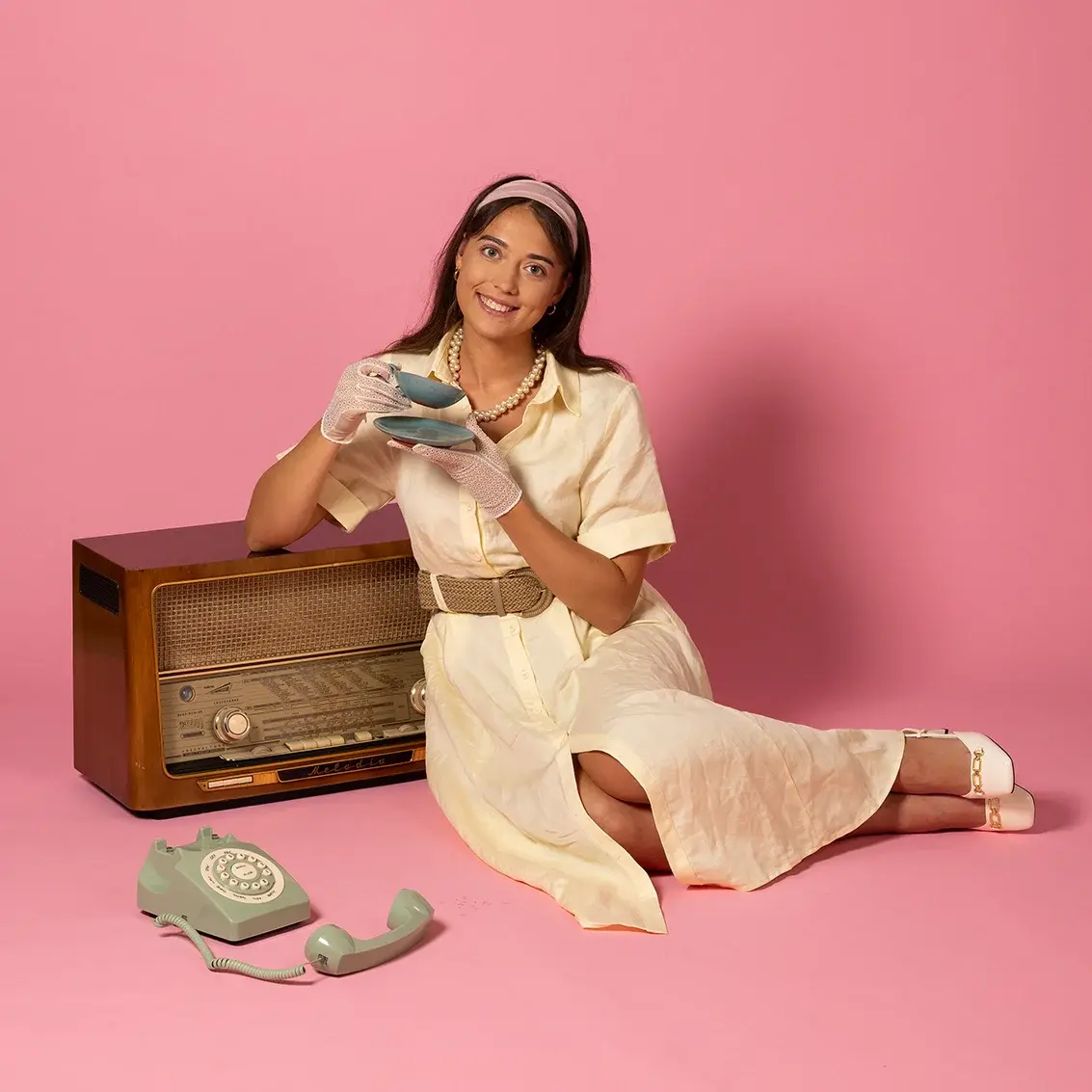 A young woman dressed in the style of the 60s is sitting on the floor leaning against an old radio and holding a teacup and saucer in her hands.
