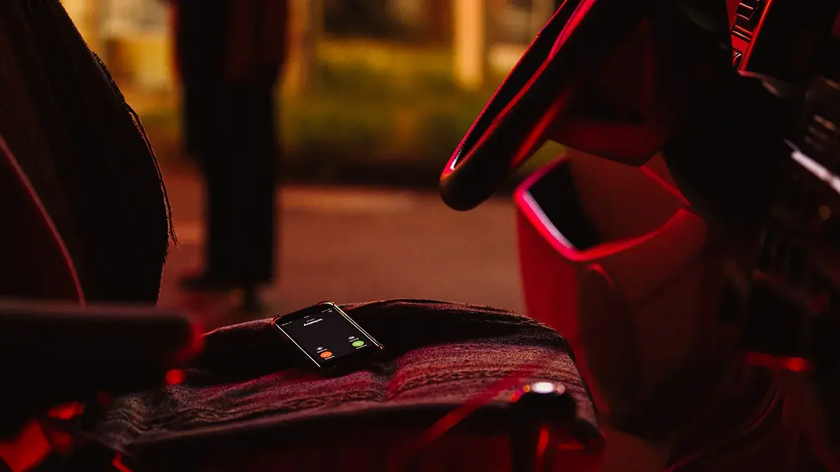 Blick auf den Fahrersitz eines Autos. Auf dem ein Smarthphone liegt. Das rötliche Licht der Tankstelle beleuchtet den Fahrzeuginnenraum.