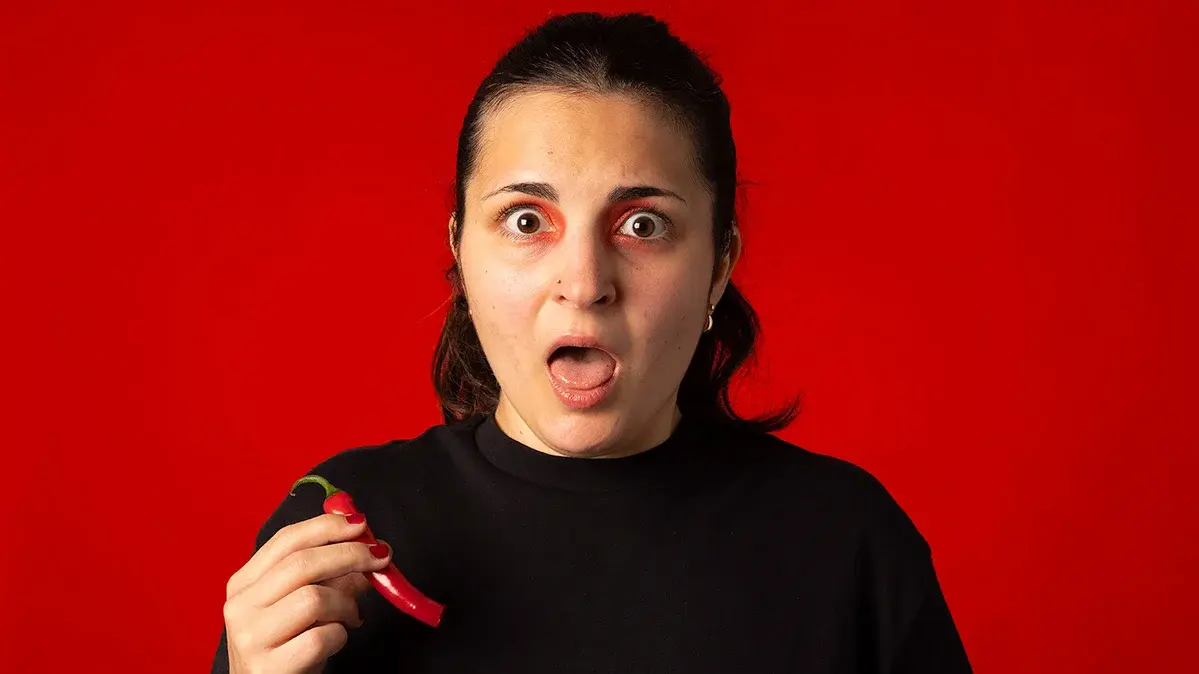 Eine junge Frau hält eine rote Pepperoni in der Hand und reißt aufgrund der Schärfe die Augen weit auf.