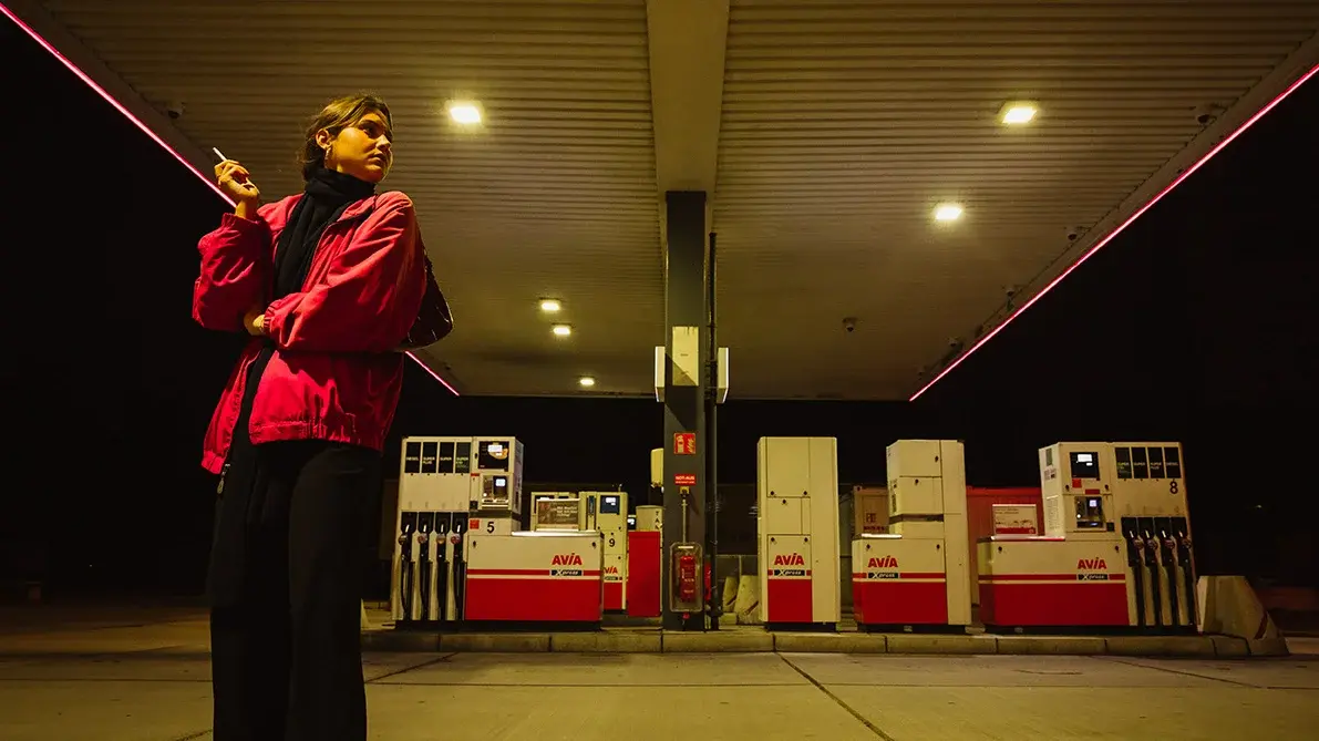Eine Frau steht in der Dunkelheit. Im Hintergrund ist eine Tankstelle zu sehen.