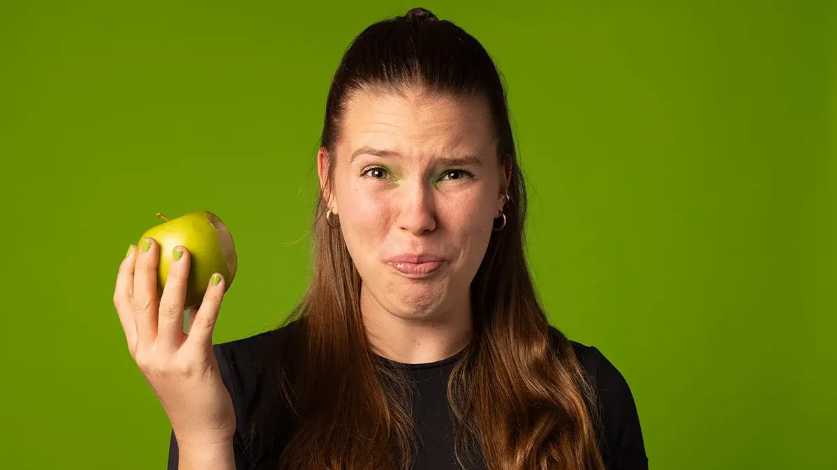 Eine junge Frau hält einen Apfel in der Hand und verzieht das Gesicht wegen dem sauren Geschmack.
