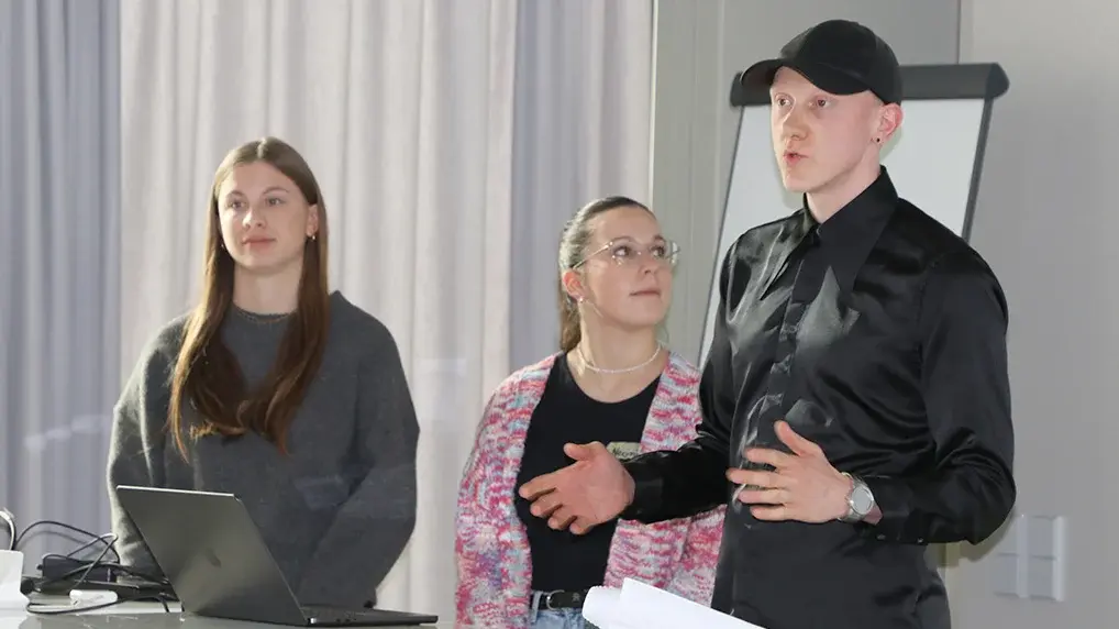 Drei Studierende stehen im Hörsaal am Pult und tragen ihre Ideen vor.