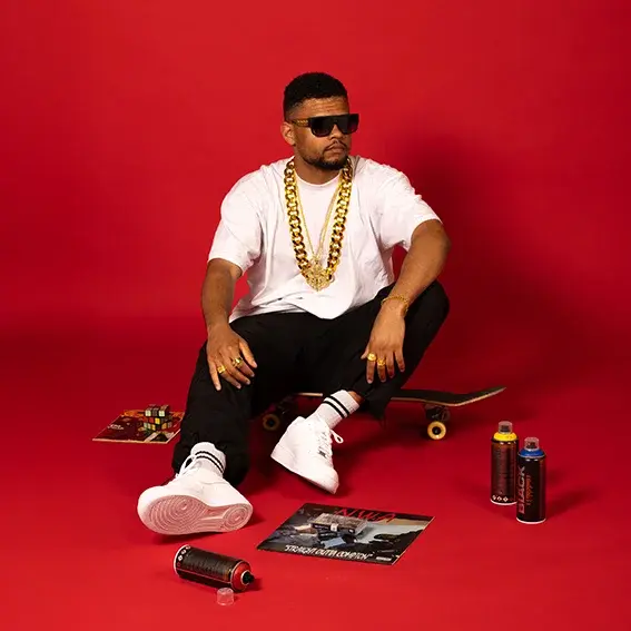 A young man dressed in 80s style is sitting on the floor. He is wearing sunglasses and a heavy gold chain.