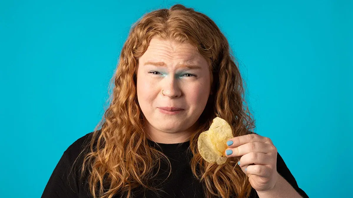 Eine junge Frau hält einen Kartoffelchip in der Hand und verzieht das Gesicht wegen dem salzigen Geschnmack.