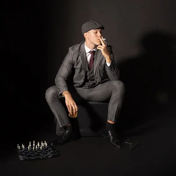 A darkly dressed young man in the style of the 1920s sits on a chair and smokes.